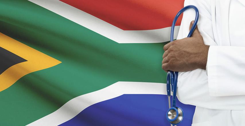 Doctor in a white coat holding a blue stethoscope beside the South African flag in the background