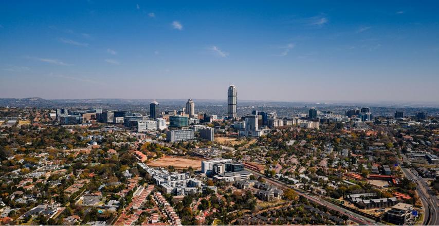 Aerial view of a city skyline with a tall central skyscraper, surrounded by dense neighborhoods and low-rise buildings under a clear blue sky.